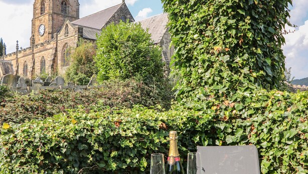 Une table avec une bouteille de vin et des verres, entourée de murs couverts de lierre et, au loin, une église.