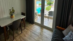 Ett rum med ett matbord och stolar, en soffa med ett mönstrat kuddöverdrag och en skjutdörr i glas som leder ut till ett utomhusområde med en pool och palmer.