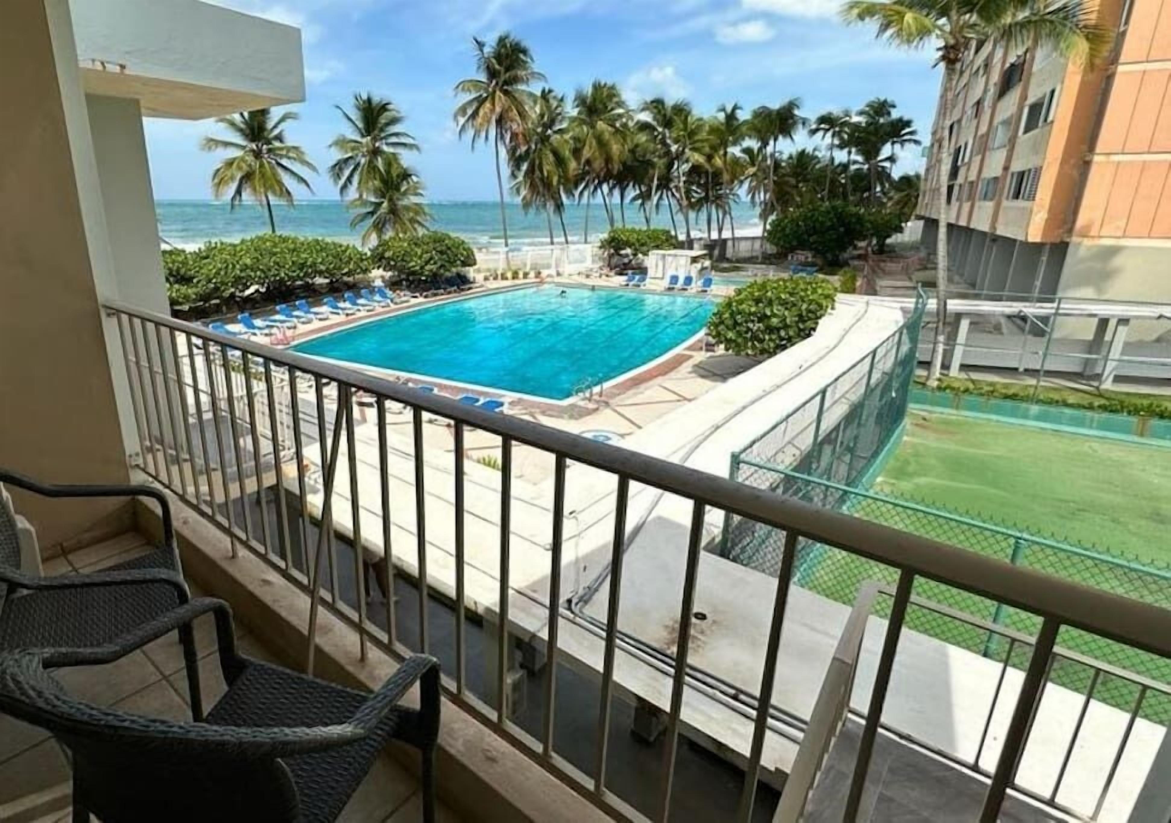 A balcony with a pool, palm trees, and a view of the ocean.