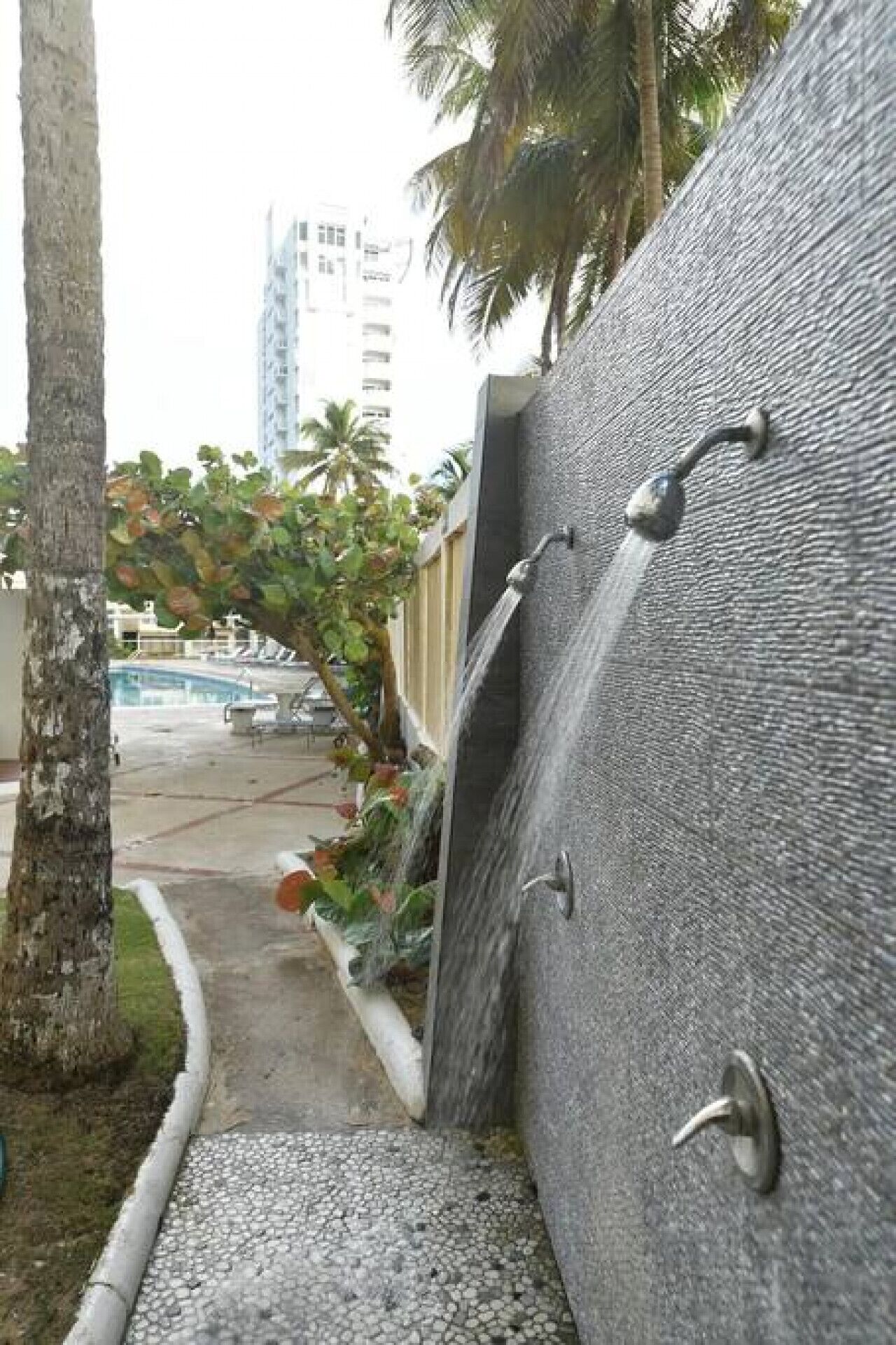 A modern building with a textured facade, a pool area with palm trees, and a staircase leading to the entrance.