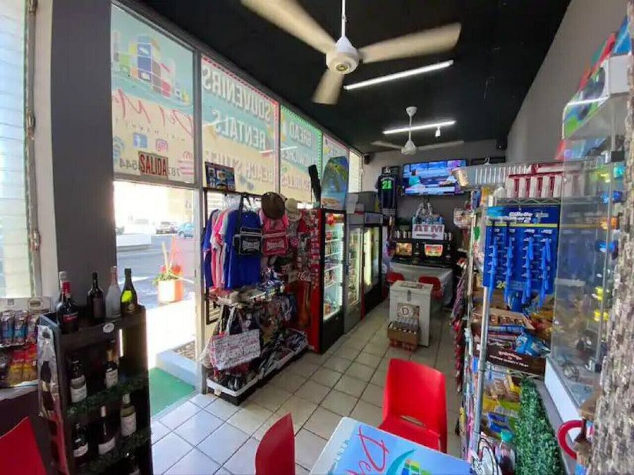 A small convenience store with a variety of products, including snacks, drinks, and household items. There are ceiling fans, a refrigerator, and a display of bottled water.