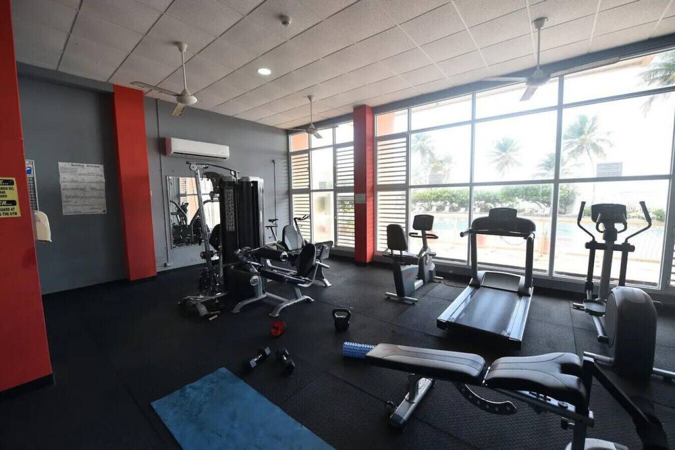 Un gimnasio con equipo de ejercicio, ventanas amplias y una pared roja.