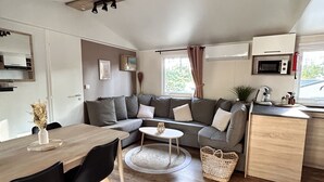 Uma sala de estar e jantar moderna, com layout aberto, teto de madeira, sofás cinza, mesa de jantar com cadeiras, uma pequena mesa de centro redonda e uma área de cozinha com micro-ondas e pia.