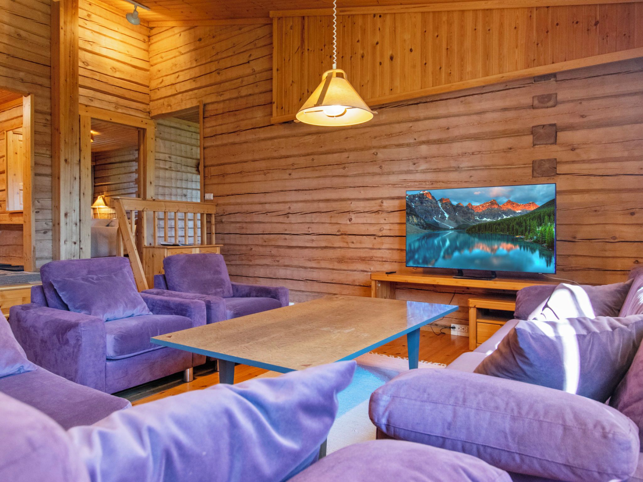 L’intérieur chaleureux d’un chalet avec des murs en bois, une grande fenêtre donnant sur un paysage montagneux, des meubles recouverts d’un tissu violet et un luminaire suspendu.