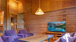 A cozy cabin interior with wooden walls, a sofa, a table, and a TV displaying a mountain landscape.