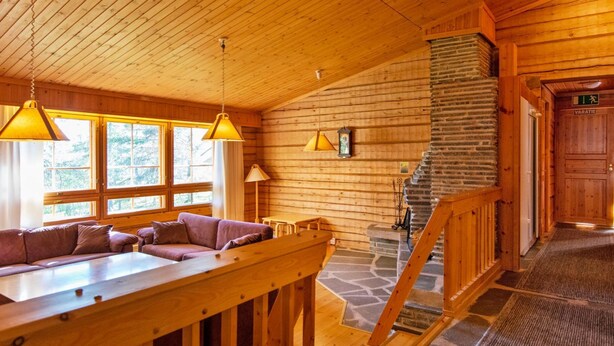 A wooden cabin interior with a stone fireplace, wooden staircase, and a sofa by a window.