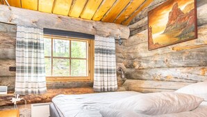 A log cabin room with a bed, a window with curtains, a painting on the wall, and a heater.