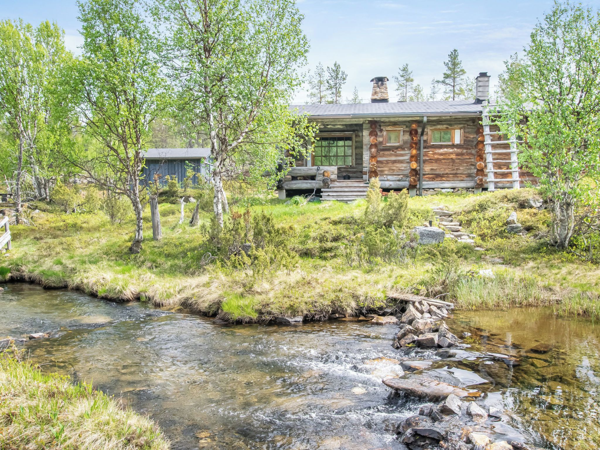 A rustic cabin with a stone chimney, a wooden deck, and a stream in front.