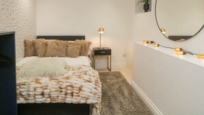 A bedroom with a white brick wall, a bed with a fluffy blanket, a bedside table with a lamp, and a mirror on the wall.