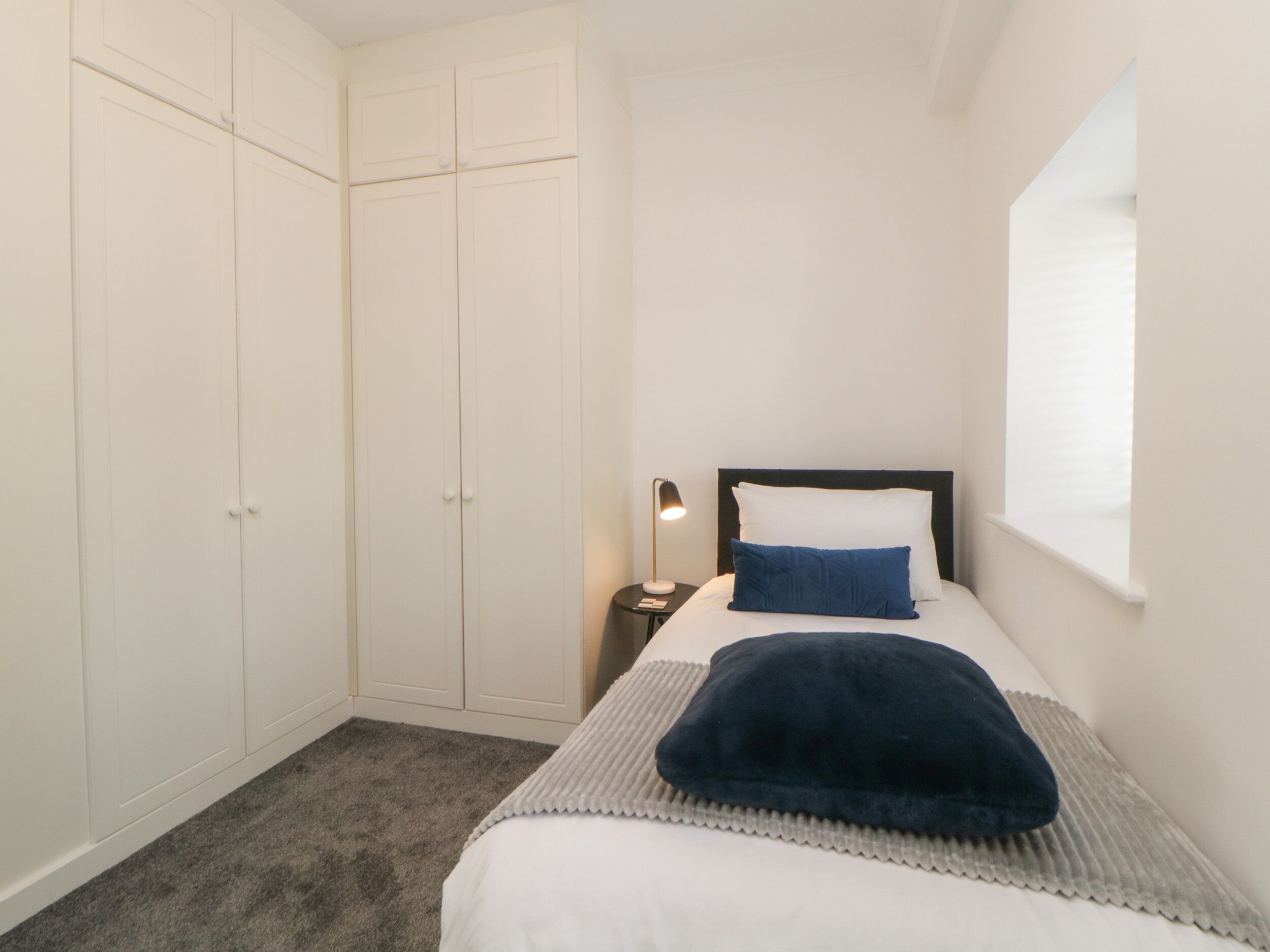 Ein Schlafzimmer mit Bett, Nachttisch, Lampe, Fenster und Kleiderschrank.
