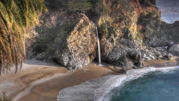 A beach with a waterfall, rocky cliffs, and ocean waves.