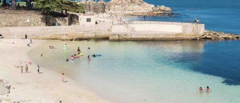 A beach with clear water, rocky outcrops, and a few people swimming and walking.