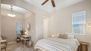 Une chambre à coucher avec un grand lit, un bureau avec une chaise, un ventilateur de plafond et une fenêtre avec des stores.