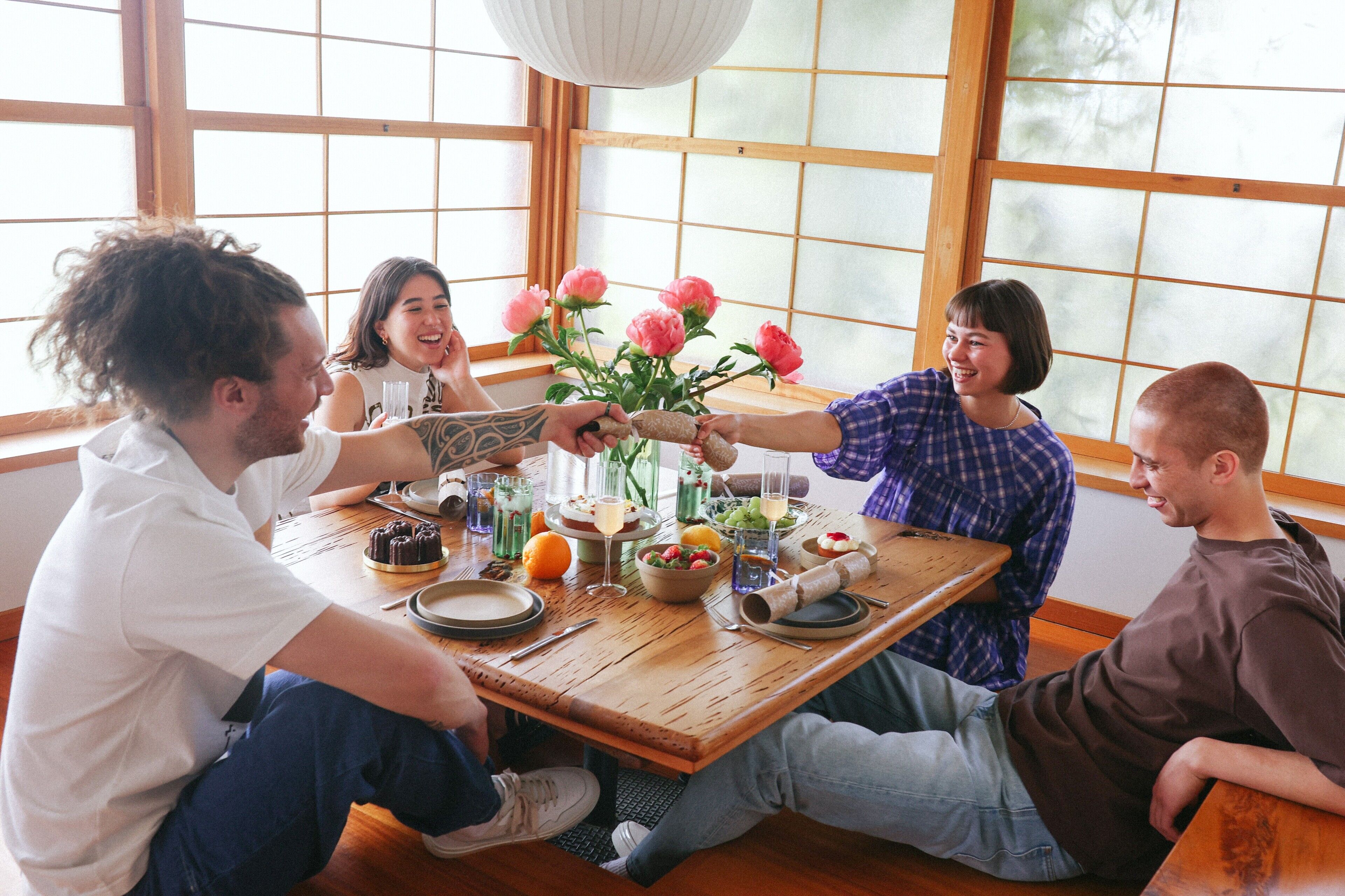 A group of people sitting around a wooden table with food and drinks, in a room with large windows and a hanging light fixture.