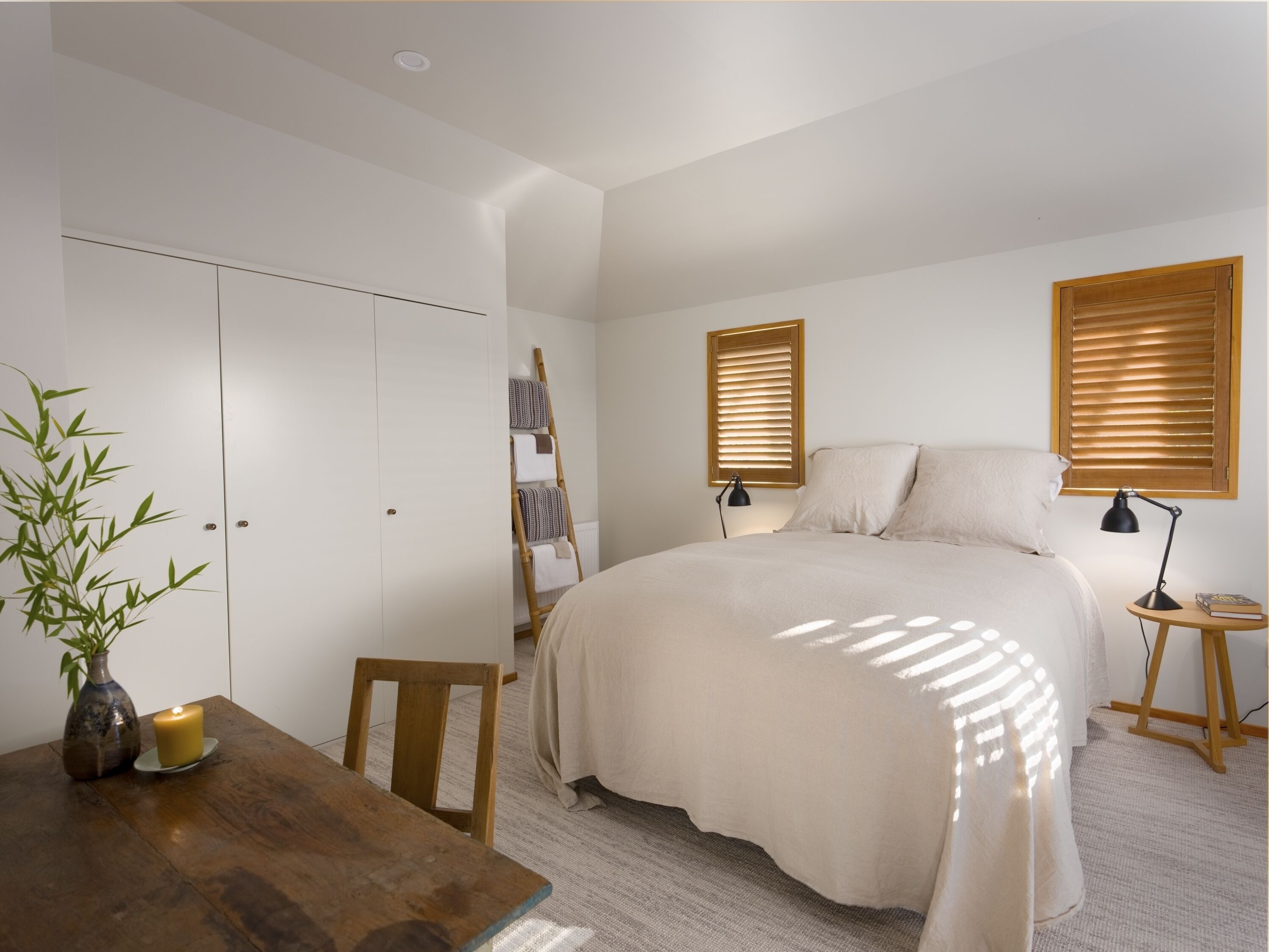 A bedroom with a bed, bedside tables, a lamp, a window with blinds, a wardrobe, and a small table with a candle.