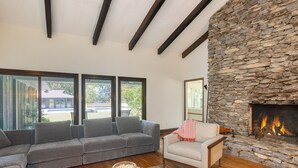 Ein Wohnzimmer mit einem grauen Ecksofa, einem weißen Sessel, einem Kamin aus Naturstein und einem großen Fenster mit Blick auf eine Terrasse.