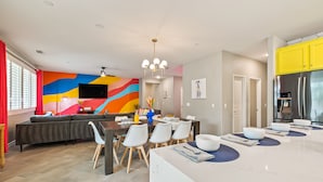 Uma cozinha moderna com uma ilha grande, uma sala de jantar com um mural colorido e uma sala de estar com sofá e televisão.