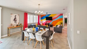 Uma área de refeições moderna com uma mesa de madeira escura, cadeiras brancas e um mural abstrato colorido ao fundo.