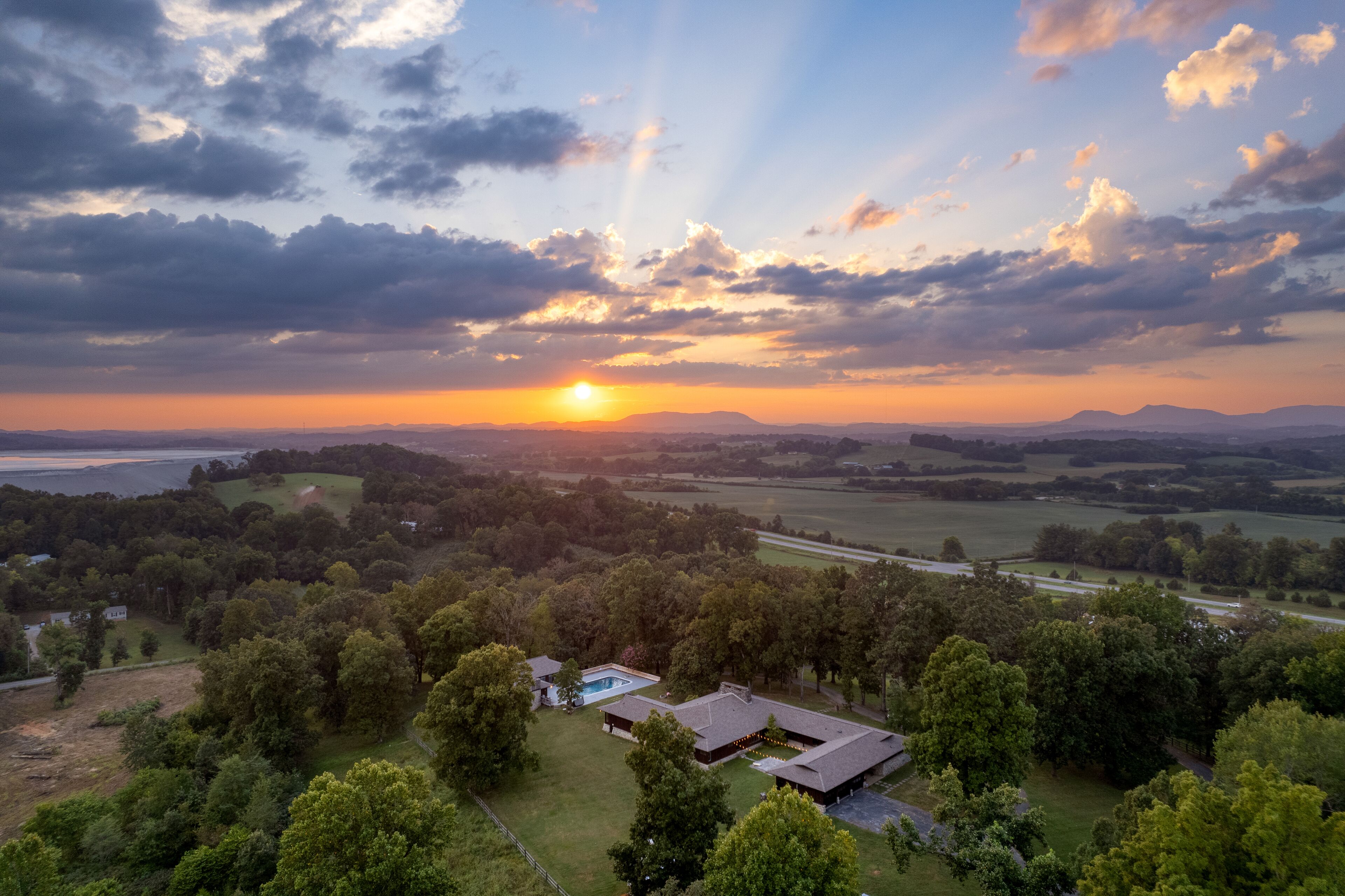 A large house with a swimming pool, surrounded by lush greenery and a scenic view of the sunset over a valley.