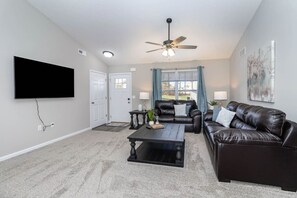 A living room with a flat-screen TV, a ceiling fan, a coffee table, two leather sofas, and a window with curtains.
