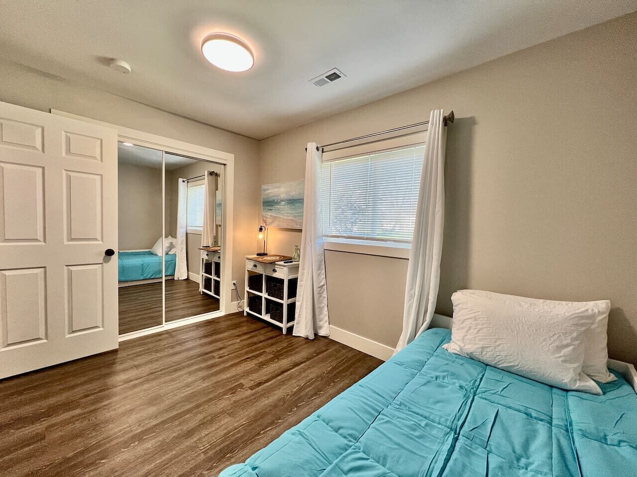 A bedroom with a bed, a nightstand, a window with curtains, a door leading to another room, and a ceiling light.