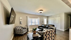 A living room with a sofa, ottoman, and coffee table, a flat-screen TV mounted on the wall, and a window with curtains.