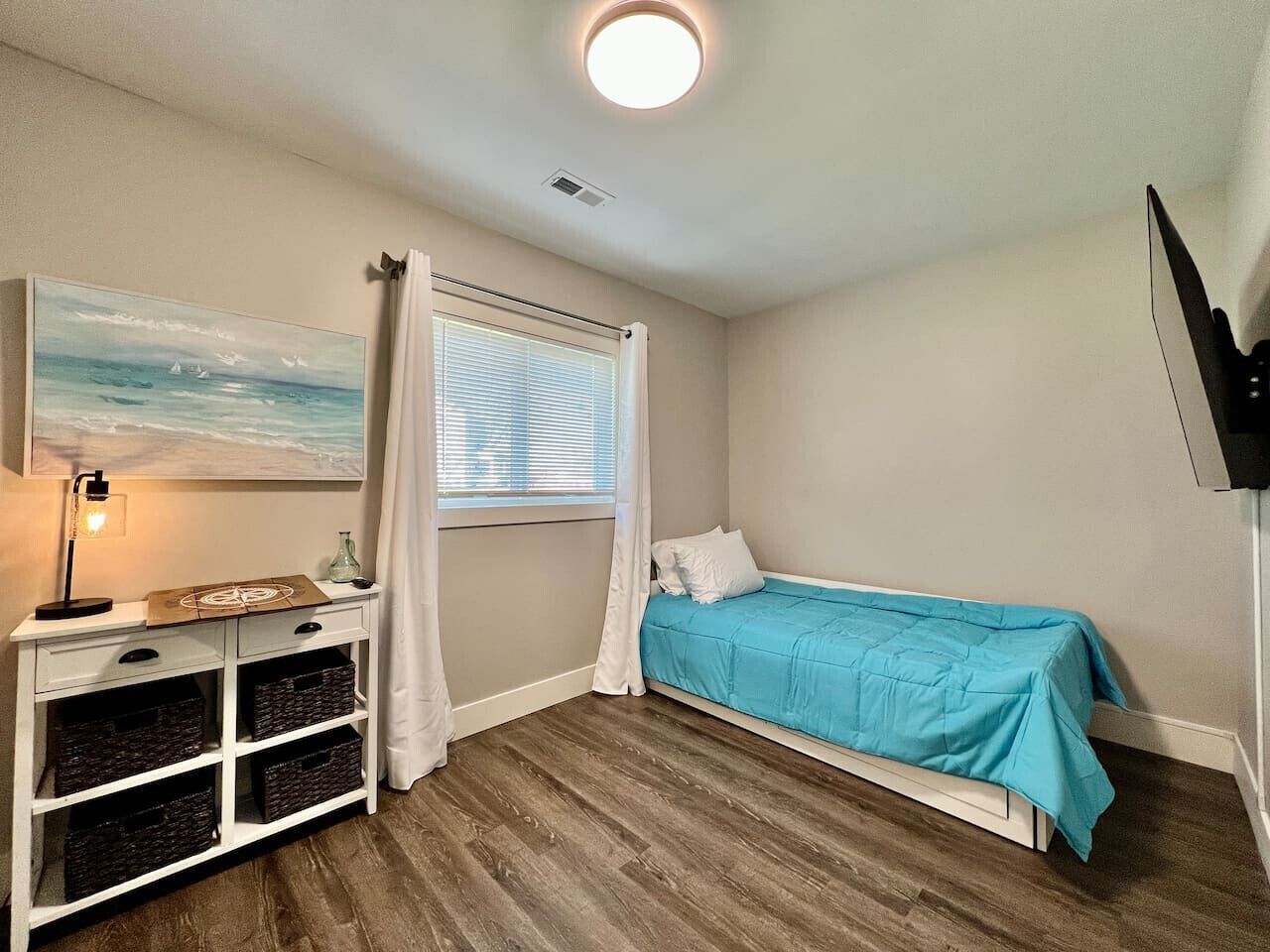 Quarto com cama, escrivaninha com espaço de armazenamento, um quadro e janela com cortinas.