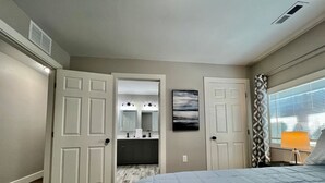A bedroom with a bed, a nightstand, a lamp, a painting, and a bathroom visible through an open door.