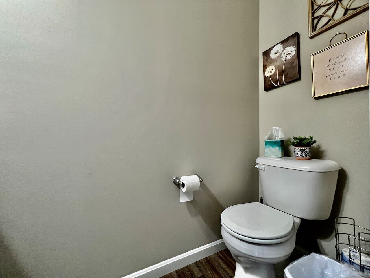 Une salle de bain avec des toilettes, un rouleau de papier toilette, une petite plante et une citation encadrée accrochée au mur.
