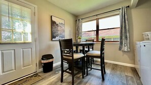 Una cocina con una mesa de comedor, sillas, un cubo de basura, un refrigerador y una ventana con cortinas.