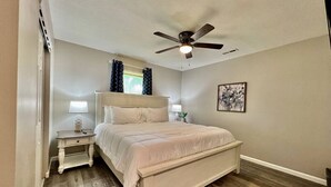Un dormitorio con cama, mesitas de noche, ventilador de techo y un cuadro en la pared.