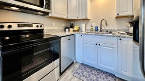 Una cocina con armarios blancos, un microondas de acero inoxidable, un horno negro y un lavamanos con grifo.