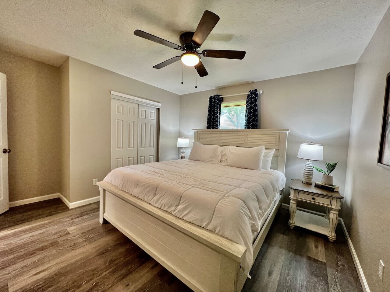 A bedroom with a bed, a ceiling fan, a nightstand with a lamp, and a door.