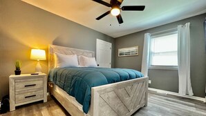 Quarto com cama, mesa de cabeceira, luminária, ventilador de teto, janela com cortinas e uma obra de arte pendurada na parede.