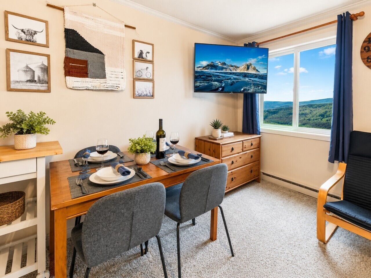A dining area with a wooden table, two chairs, a TV mounted on the wall, and a window with curtains.