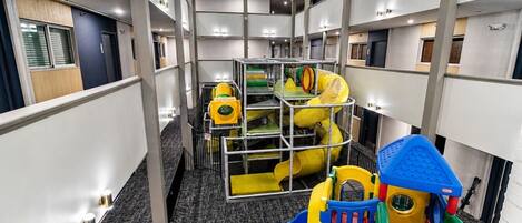 Zona de juegos infantiles con toboganes y estructuras para escalar, ubicada en un pasillo de hotel con varios pisos y diseño moderno.