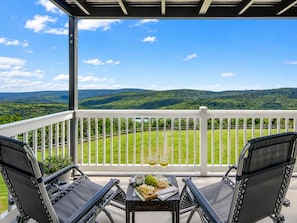 A balcony with two chairs, a table with a bottle of wine, and a view of green hills. - ML258 Slope Side Getaway with Deck (Snowshoe)