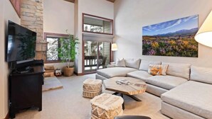 A spacious living room with a large sectional sofa, a flat-screen TV, a fireplace, and a large window offering a view of the outdoors. - Ridgepoint Townhome 142 - 3.5 Bedroom plus Loft Residence (Avon)