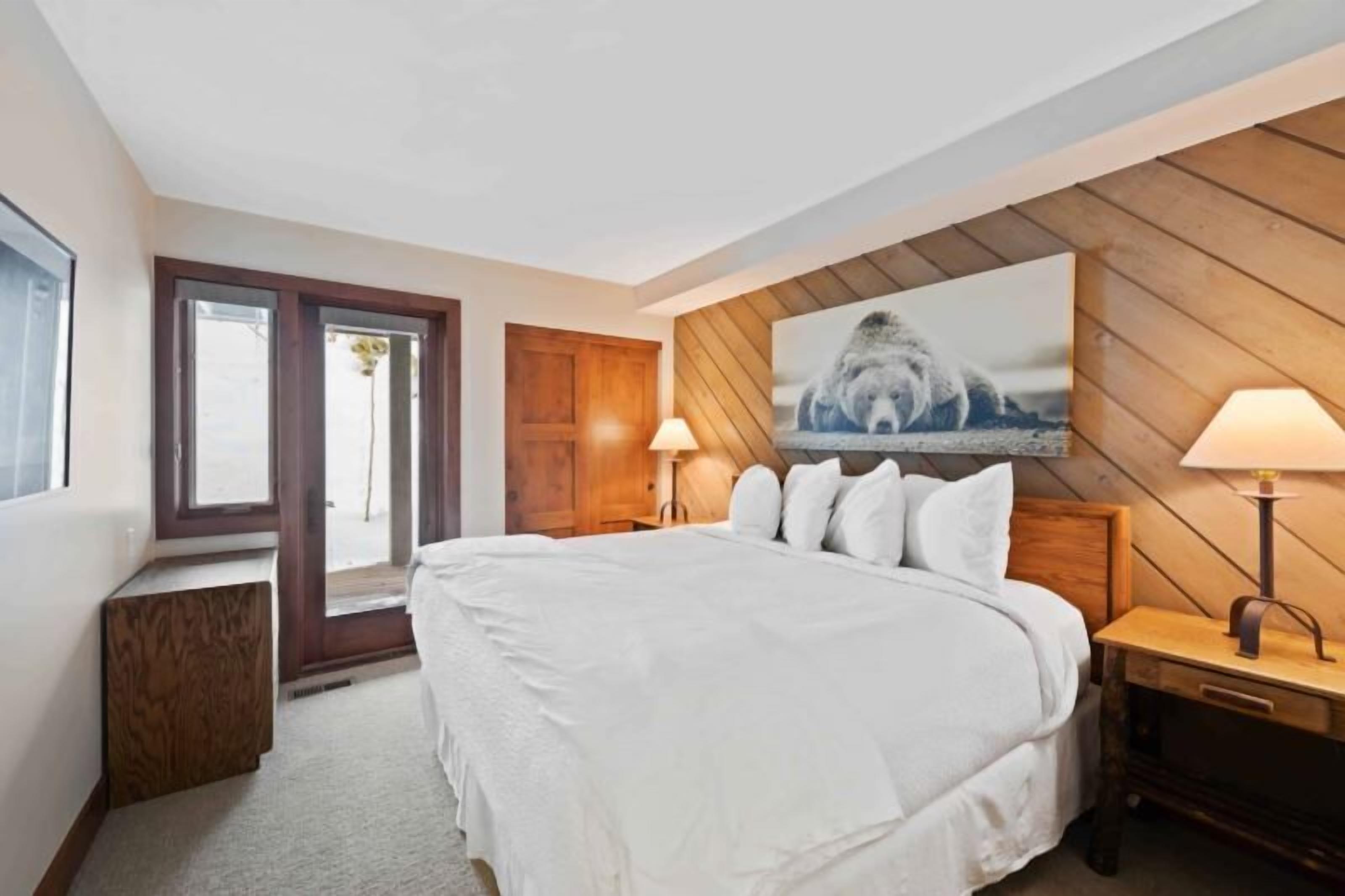 A bedroom with a large bed, wooden headboard, two bedside lamps, a wooden wall panel, and a window with a view.