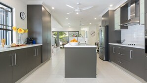 A modern kitchen with dark cabinets, a central island, and a large window. - Bella Beach House | Port Douglas (PORT DOUGLAS)