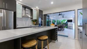 A modern kitchen with stainless steel appliances, a large island with stools, and a view into a dining area with a ceiling fan. - Bella Beach House | Port Douglas (PORT DOUGLAS)