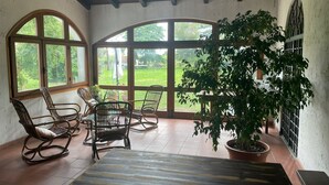 A room with large arched windows, wicker chairs, a potted plant, and a tiled floor.