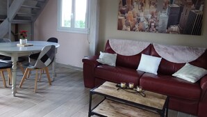A living room with a red leather sofa, a coffee table with candles, a dining table with chairs, and a cityscape painting on the wall. - Holiday Home 'Gite En Baie De Somme' with Private Terrace, Private Garden and Wi-Fi (Saint-Quentin-en-Tourmont)