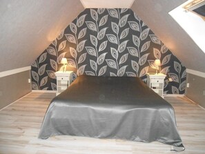 Camera da letto con soffitto spiovente, un letto con coperta grigia, due comodini con lampade e carta da parati fantasia.