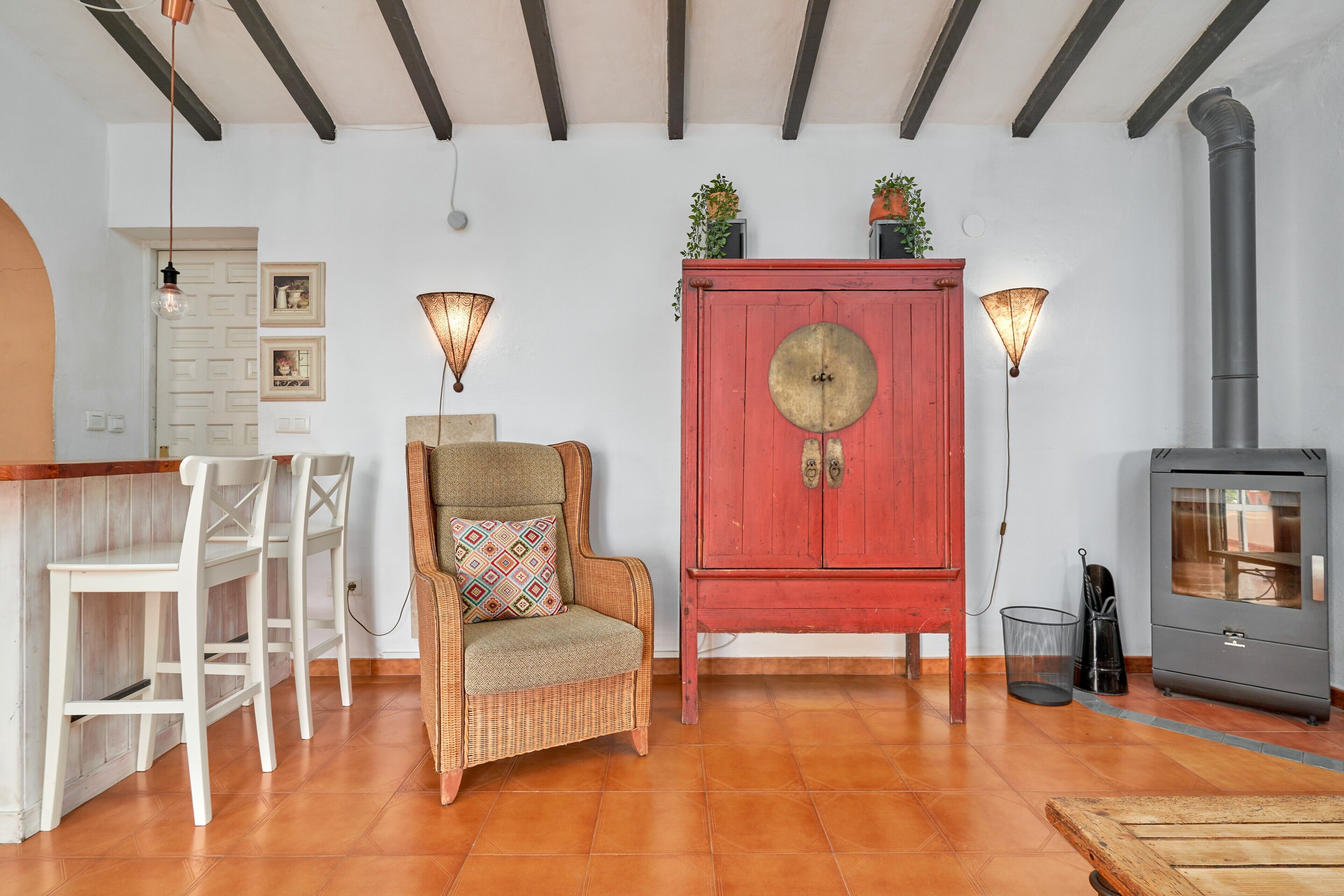 A cozy room with a wooden floor, a red cabinet, a wicker chair, white chairs, a small table, and a wood-burning stove.