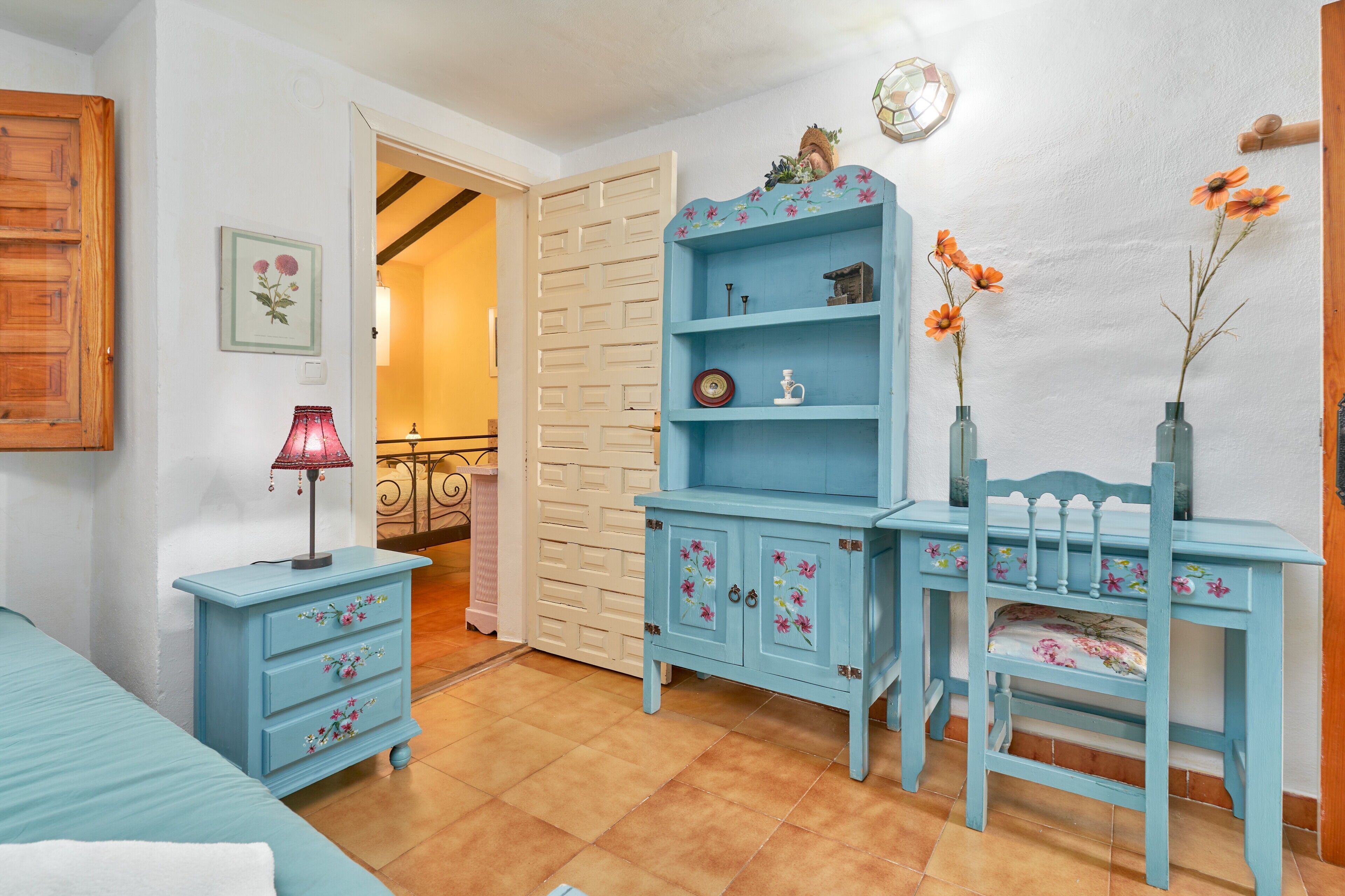A room with a bed, a dresser, a chair, and a desk with a vase of flowers.