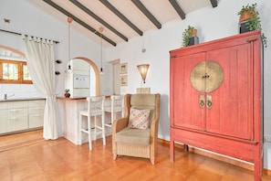A room with a wicker chair, a red cabinet, a white chair, a kitchen area, and a window with curtains.