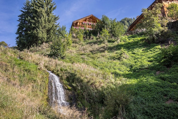 A wooden cabin with a waterfall in the foreground, surrounded by lush greenery and trees.