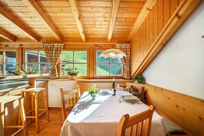 Ein Holzblockhaus-Interieur mit einem Esstisch, gedeckt für zwei Personen, Holzstühle, einem Fenster mit Vorhängen und Blick ins Grüne.