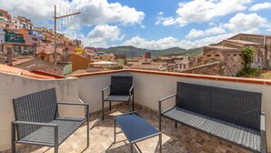 Une terrasse sur le toit, avec deux chaises et une table, d’où l’on peut admirer un paysage urbain avec des bâtiments aux couleurs vives et des montagnes à l’horizon.